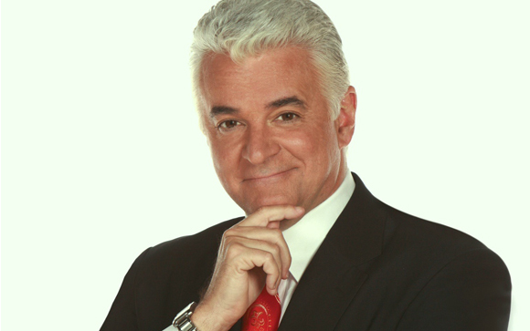 A man with silver hair wearing a black suit and red tie, resting his chin on his hand.