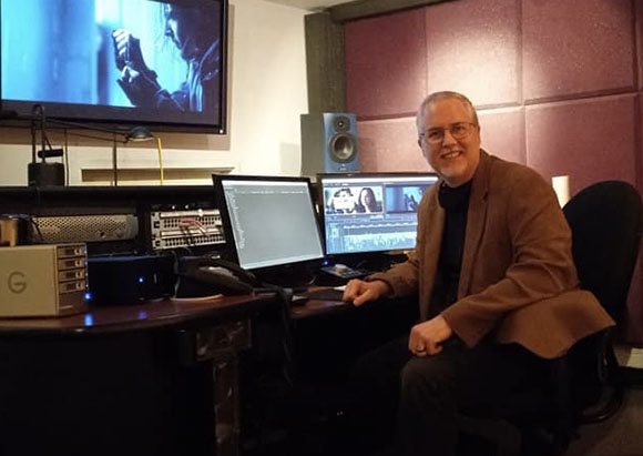 A man in a brown blazer sits at a desk with two computer monitors and a large screen in a studio.