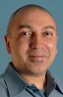 A smiling man with short hair and a small goatee wearing a blue collared shirt against a blue background.