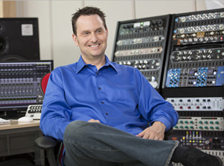 A smiling man in a blue button-down shirt sits in a chair surrounded by audio mixing equipment.
