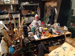 A man with white hair and a beard sits behind a workbench filled with tools and supplies in a crowded room.