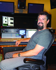 A man with a beard sits in a chair in front of a large audio mixing console and multiple monitors.