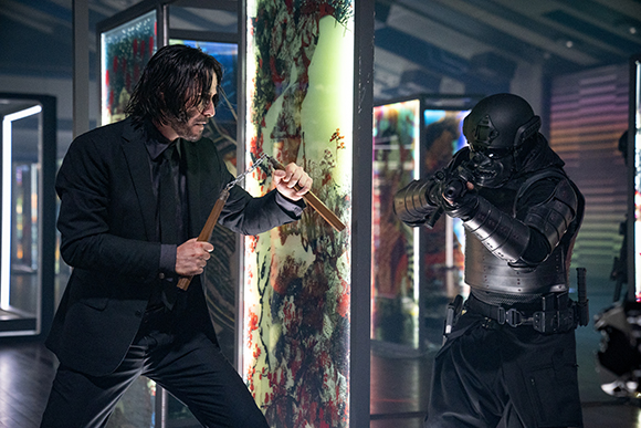 A man in a black suit uses a wooden tool to fight an armored soldier in a dark room with glowing displays.