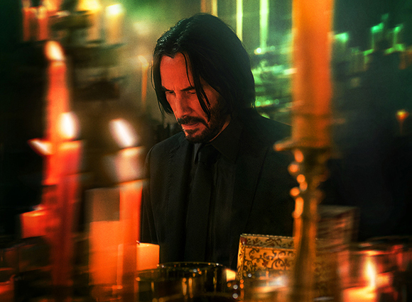 A man with long dark hair and a beard wearing a black suit stands behind several glowing orange candles.