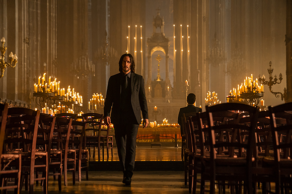 A man in a black suit walks down an aisle in a dark, candlelit cathedral.