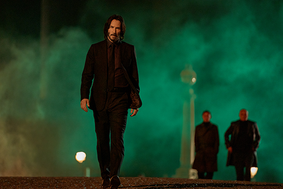 A man in a dark suit walks toward the camera through thick green smoke at night.