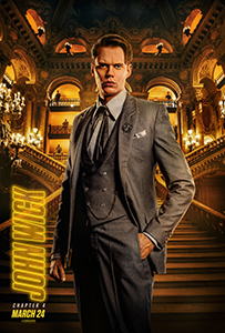 A man in a grey suit stands before a grand, golden-lit staircase and arched balconies.