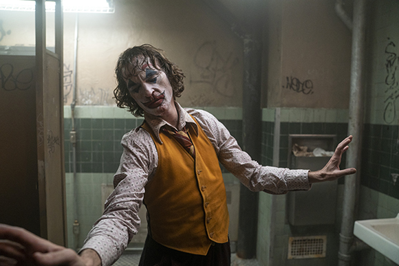 A man with clown makeup and a yellow vest dances in a grimy, graffiti-covered bathroom.