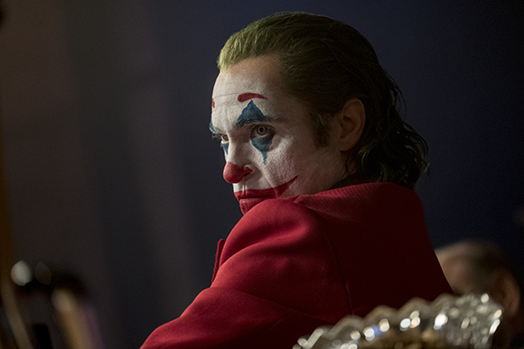 A man with white, blue, and red clown makeup wearing a red suit looks back over his shoulder.