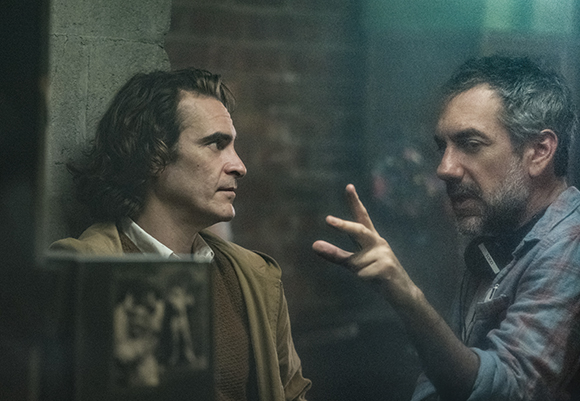 A man with wavy hair looks toward a bearded man gesturing with his hand in a dimly lit setting.