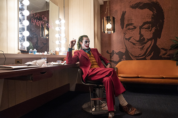 A man in clown makeup and a red suit sits in a chair in a dressing room next to a lit vanity mirror.