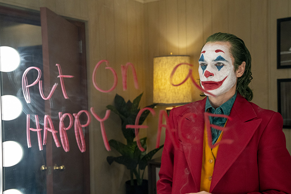 A man in clown makeup and a red suit looks through glass with 'Put on a HAPPY FACE' written in pink and white.