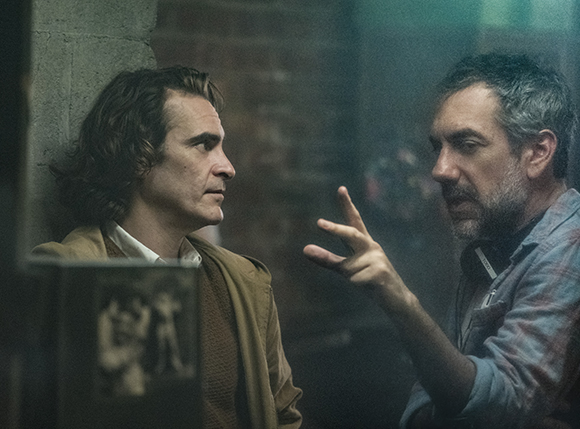 A man with wavy hair looks at another man who is gesturing with his hand during a conversation.