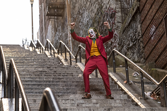 A man in a red suit and clown makeup dances with raised arms on a long outdoor concrete staircase.