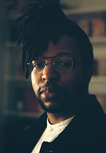 A man with dark dreadlocks and glasses looks directly at the camera in a dimly lit setting.