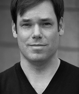 Black and white headshot of a man with short dark hair wearing a dark V-neck shirt.