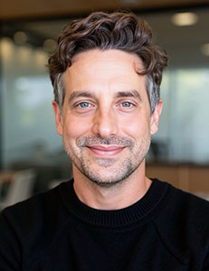 A man with wavy brown hair and light eyes smiles at the camera in a headshot.