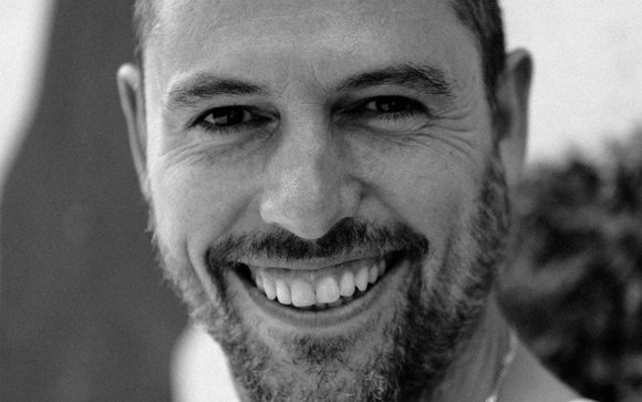 Close-up black and white portrait of a man smiling with visible teeth and a beard.
