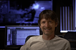 A man with brown hair and a light-colored shirt smiles in a dark studio with glowing monitors.