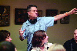 A man in a light blue shirt gestures with his arms wide in a room with framed art on the wall.
