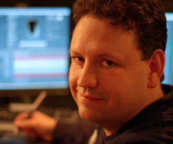 A man with curly hair smiling slightly in front of computer monitors in a dimly lit room.