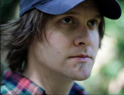 Close-up of a man wearing a blue baseball cap and a red and blue plaid shirt, looking upwards with a thoughtful expression. T