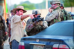 A man in a tan hat points while soldiers in camouflage gear stand behind a blue Honda sedan.