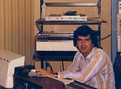 A man in a striped shirt sits at a desk in a room filled with electronic equipment and shelving.