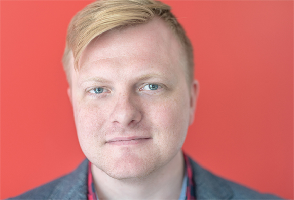 Close-up portrait of a man with light blond hair and blue eyes against a solid red background.
