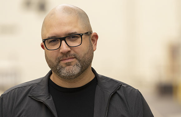 A bald man with dark-rimmed glasses and a salt-and-pepper beard wearing a black jacket.