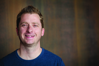A smiling man with short blond hair wearing a blue crewneck t-shirt against a dark background.