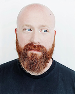 A bald man with a thick red beard and blue eyes looking off-camera against a white background.