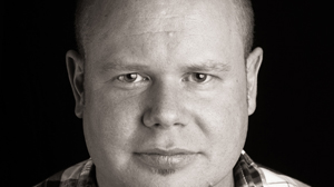 Close-up black and white portrait of a man looking directly at the camera against a dark background.