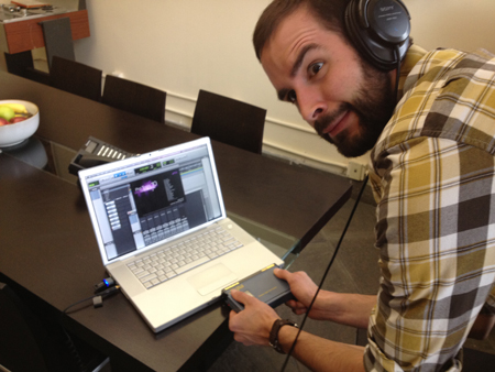 A man in a plaid shirt wearing headphones and using a laptop and audio interface at a dark table.