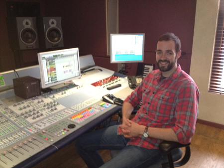 A man in a red plaid shirt sits at a professional audio mixing desk with computer monitors.