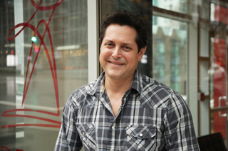 A smiling man with dark hair wearing a grey and white plaid button-down shirt.