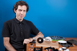 A man in a black shirt sits at a wooden table with film reels, a film viewer, and other related equipment.