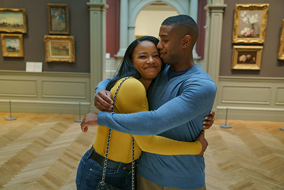 A man in a blue shirt and a woman in a yellow shirt embrace in a museum gallery surrounded by framed art.