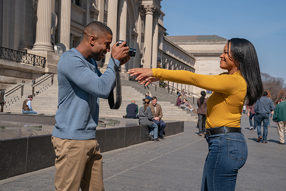 A man in a blue sweater takes a photo of a smiling woman in a yellow shirt outdoors.