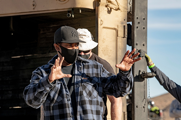 A man in a black cap and plaid shirt gestures with open palms toward a large vehicle.