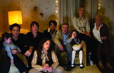 A diverse group of adults and a child sitting together in a dimly lit living room with surprised expressions.
