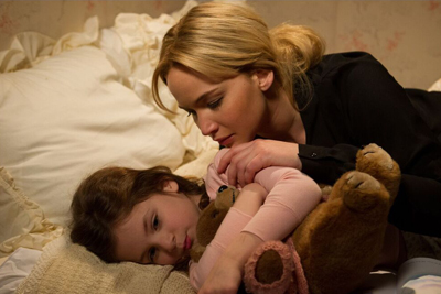 A blonde woman leans over a young girl lying in bed holding a stuffed animal.