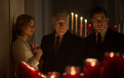 A woman with hands clasped and two men in dark clothing stand before glowing red candles in a dim room.