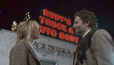 A man and woman smile at each other at night in front of a glowing Rudy's Truck & Auto Body sign.