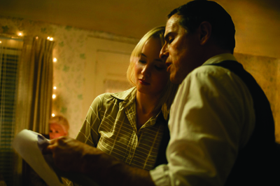 A man in a white shirt and vest and a blonde woman in a plaid shirt look down at a piece of paper.