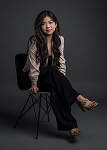 A woman with long dark hair sits on a black chair wearing a light blouse and black trousers.