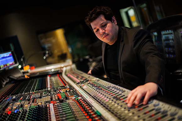A man in a dark suit adjusts sliders on a large professional audio mixing console.