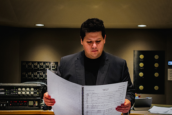 A man in a dark blazer looks down while holding open a large book of musical scores in a studio.