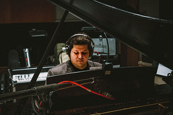 A man with headphones sits behind a black grand piano in a recording studio.