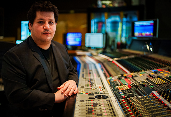 A man in a black blazer sits behind a large, complex professional audio mixing console in a dark studio.
